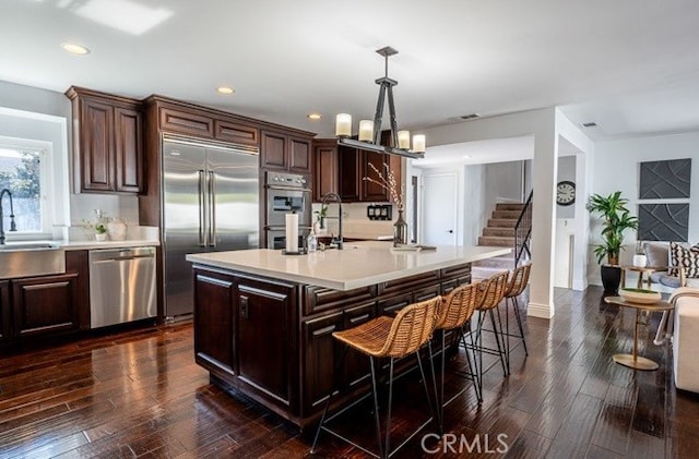 kitchen with sink, appliances with stainless steel finishes, hanging light fixtures, dark brown cabinetry, and an island with sink