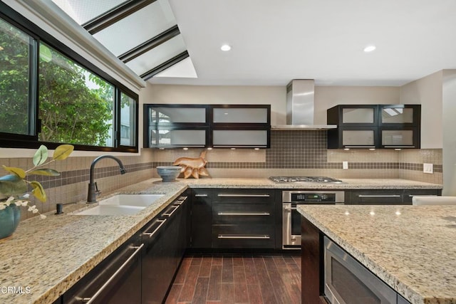 kitchen featuring light stone countertops, a skylight, wall chimney exhaust hood, stainless steel appliances, and sink