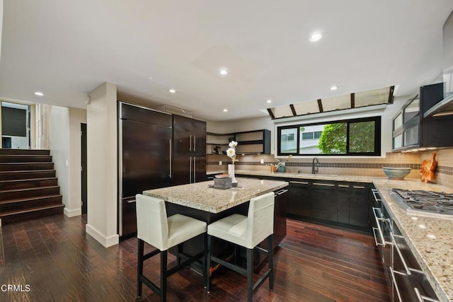 kitchen featuring light stone countertops, a center island, stainless steel gas cooktop, built in refrigerator, and a kitchen bar