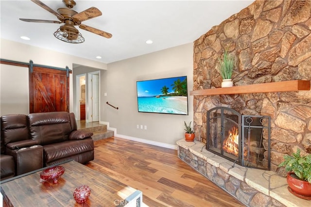 living room featuring ceiling fan, a barn door, wood-type flooring, and a stone fireplace