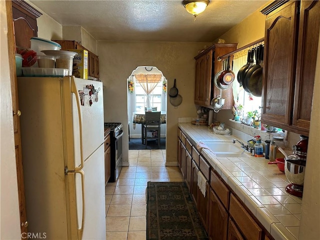 kitchen with tile counters, white refrigerator, sink, gas range, and light tile patterned floors