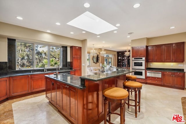 kitchen with a skylight, dark stone countertops, an island with sink, multiple ovens, and a breakfast bar area