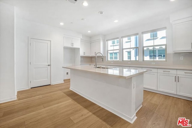 kitchen with white cabinets, light wood-type flooring, a kitchen island with sink, and sink