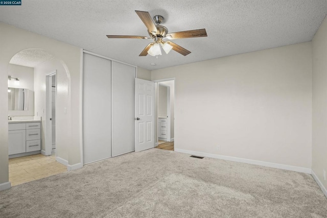 unfurnished bedroom featuring ceiling fan, ensuite bathroom, a textured ceiling, light carpet, and a closet