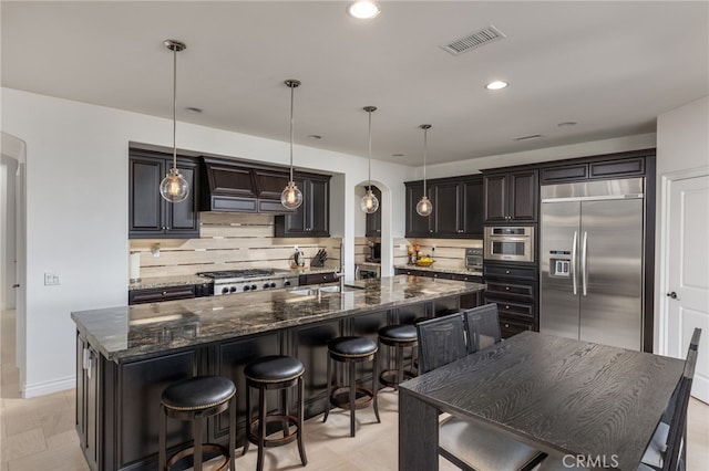 kitchen with tasteful backsplash, dark stone countertops, hanging light fixtures, an island with sink, and stainless steel appliances