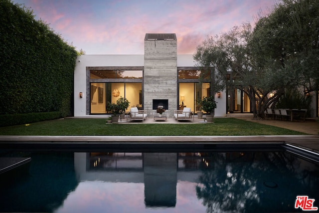 back house at dusk with a patio, an outdoor living space with a fireplace, and a lawn