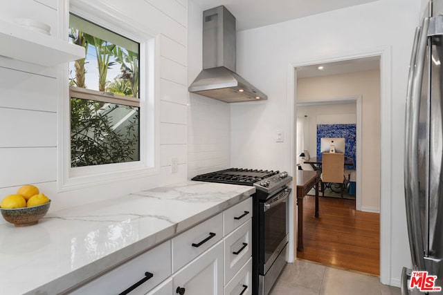 kitchen featuring light stone countertops, appliances with stainless steel finishes, wall chimney range hood, light tile patterned floors, and white cabinets