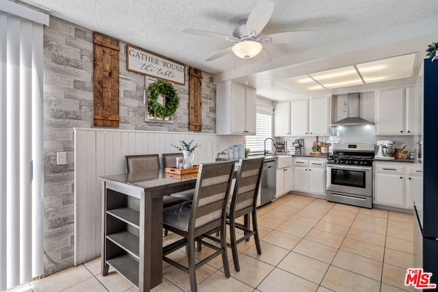 kitchen featuring stainless steel appliances, ceiling fan, wall chimney range hood, light tile patterned floors, and white cabinetry