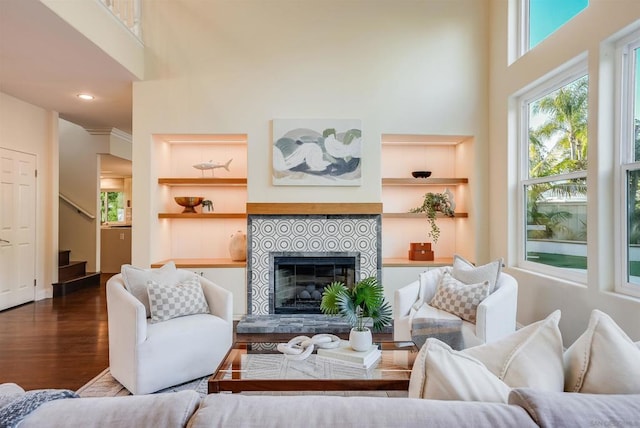 living room with a tiled fireplace, built in features, a towering ceiling, and hardwood / wood-style flooring