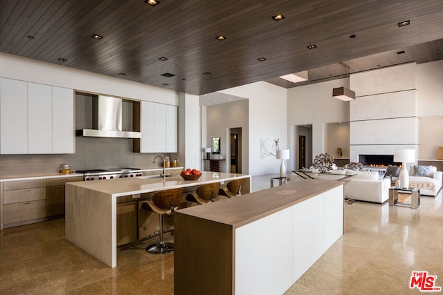 kitchen with a large island with sink, wall chimney exhaust hood, a towering ceiling, a fireplace, and white cabinetry