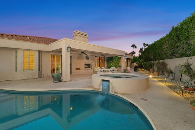 pool at dusk with an in ground hot tub, ceiling fan, a patio, and exterior fireplace