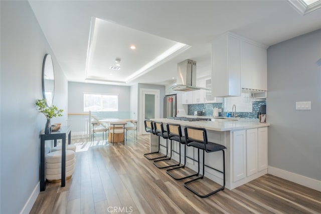 kitchen with white cabinetry, wall chimney range hood, tasteful backsplash, light hardwood / wood-style flooring, and a tray ceiling