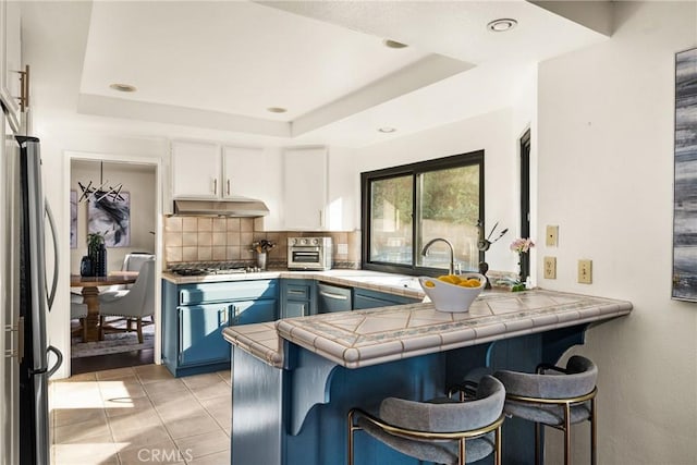 kitchen with light tile patterned flooring, blue cabinets, kitchen peninsula, a tray ceiling, and white cabinets