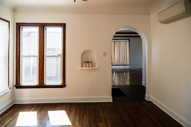 spare room featuring arched walkways, a wall mounted air conditioner, hardwood / wood-style flooring, and baseboards