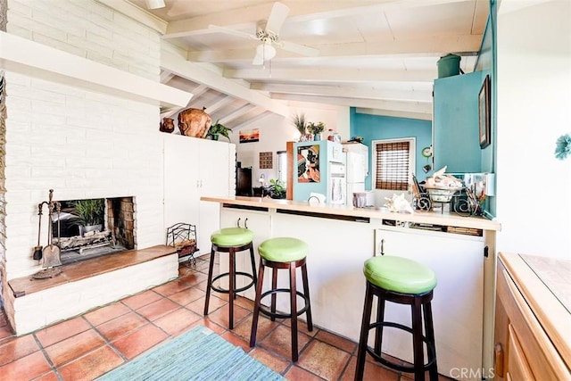 kitchen featuring ceiling fan, a brick fireplace, tile patterned flooring, lofted ceiling with beams, and a breakfast bar area