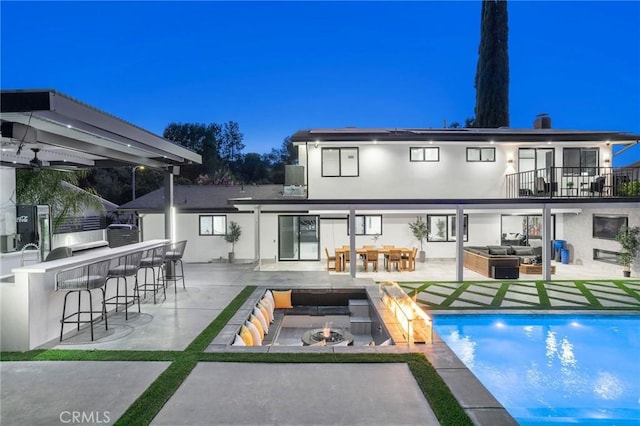 back house at dusk featuring a balcony, an outdoor bar, a patio, and an outdoor living space with a fire pit