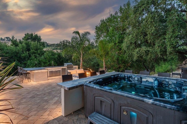 patio terrace at dusk with a bar, an outdoor kitchen, and a hot tub