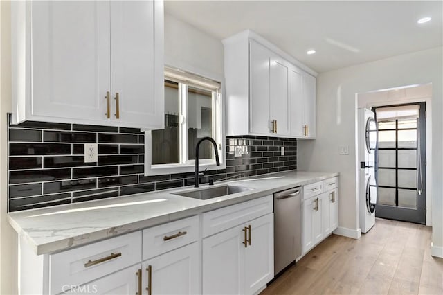 kitchen with sink, light stone counters, stainless steel dishwasher, stacked washer / dryer, and white cabinets