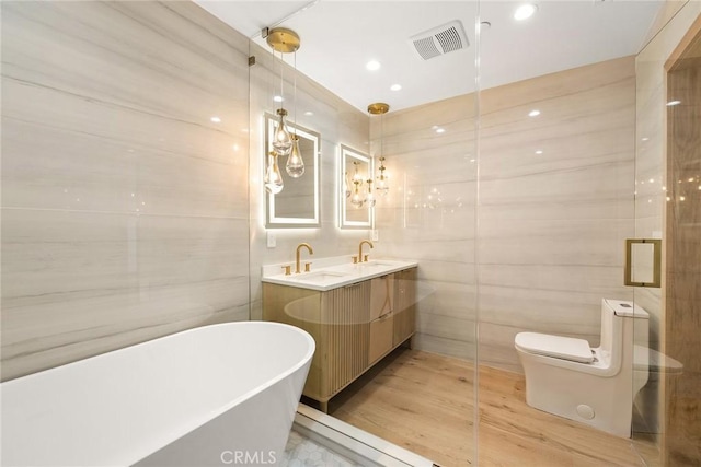 bathroom featuring toilet, visible vents, tile walls, a soaking tub, and double vanity