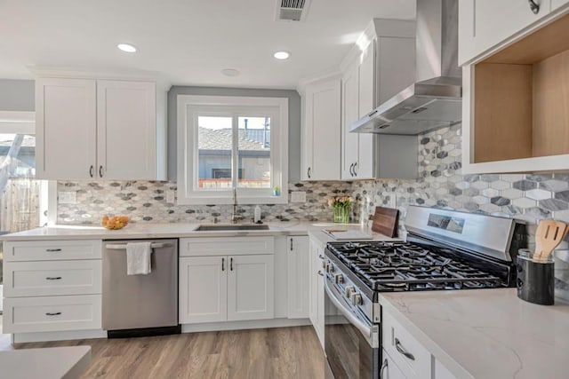 kitchen with stainless steel appliances, light stone countertops, wall chimney exhaust hood, white cabinets, and sink