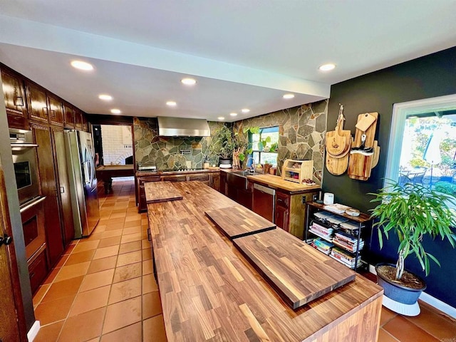 kitchen with appliances with stainless steel finishes, butcher block countertops, sink, wall chimney range hood, and light tile patterned flooring