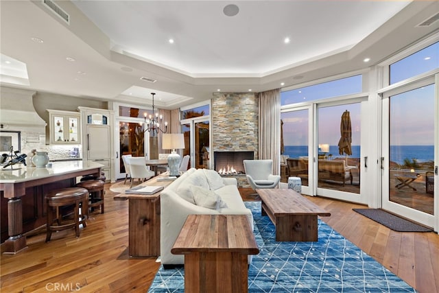 living room with a stone fireplace, an inviting chandelier, a water view, dark hardwood / wood-style floors, and a raised ceiling