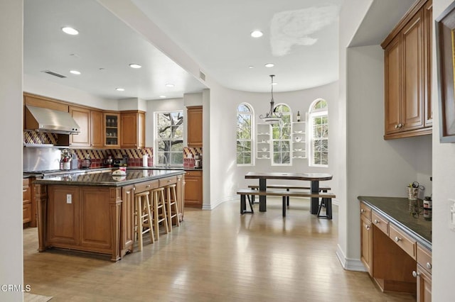 kitchen featuring dark stone counters, hanging light fixtures, a center island, wall chimney exhaust hood, and light hardwood / wood-style flooring