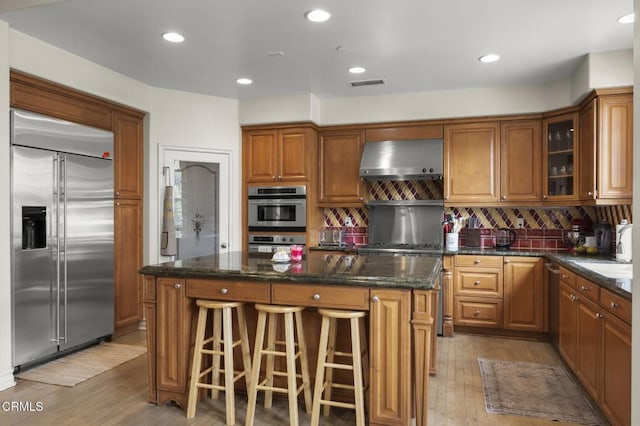 kitchen featuring wall chimney range hood, dark stone countertops, stainless steel appliances, a center island, and tasteful backsplash