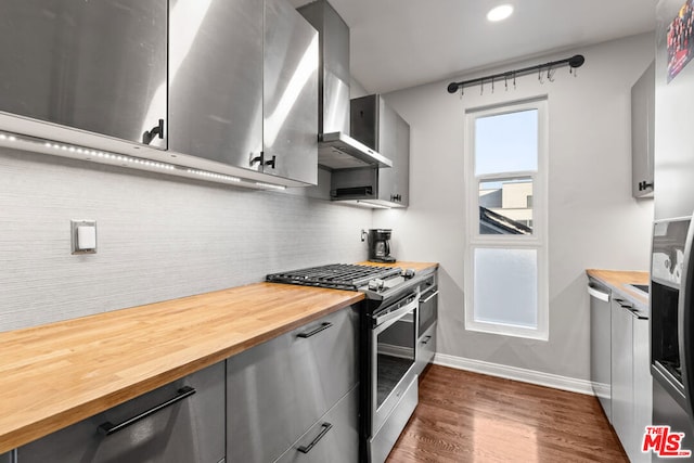 kitchen with stainless steel appliances, butcher block counters, wall chimney range hood, tasteful backsplash, and dark hardwood / wood-style flooring