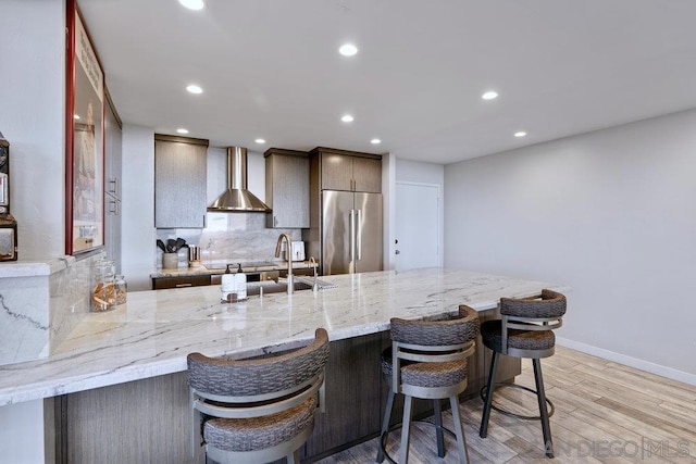 kitchen with wall chimney exhaust hood, high end fridge, light hardwood / wood-style flooring, kitchen peninsula, and a breakfast bar