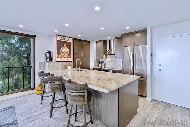 kitchen featuring kitchen peninsula, wall chimney exhaust hood, a breakfast bar, light hardwood / wood-style floors, and built in refrigerator