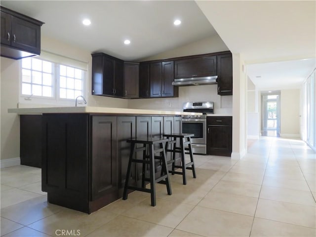 kitchen featuring stainless steel range, dark brown cabinets, vaulted ceiling, light tile patterned floors, and a breakfast bar area