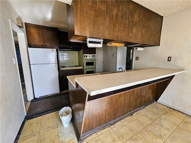 kitchen featuring kitchen peninsula, dark brown cabinetry, double oven, sink, and white refrigerator