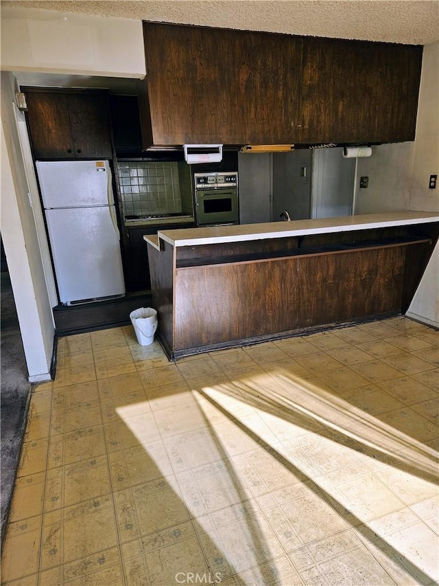 kitchen featuring oven, white refrigerator, dark brown cabinetry, and kitchen peninsula