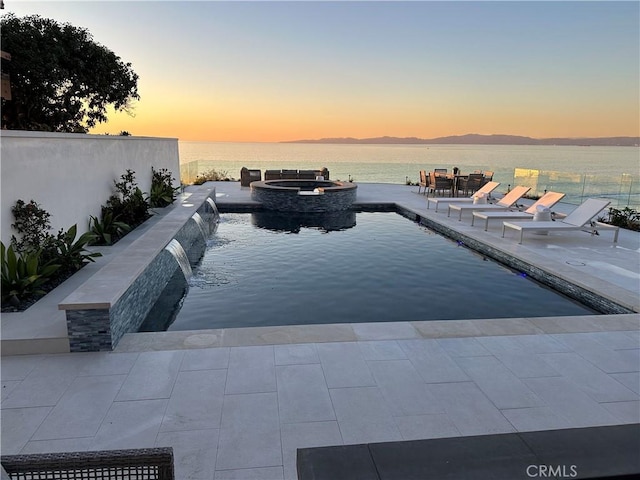 pool at dusk featuring a water view, pool water feature, and an in ground hot tub