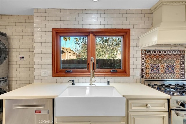 kitchen featuring stainless steel stove, cream cabinets, custom range hood, stacked washer / drying machine, and sink