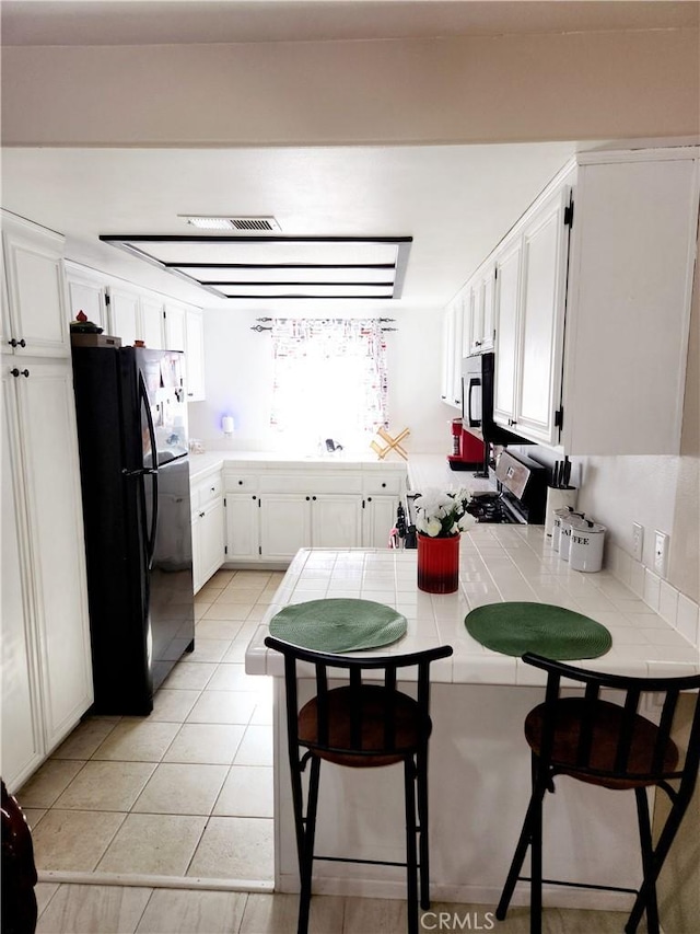 kitchen featuring a breakfast bar, kitchen peninsula, tile countertops, black refrigerator, and white cabinets