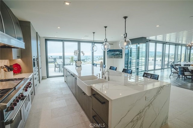 kitchen featuring decorative light fixtures, wall chimney range hood, a wall of windows, a large island with sink, and sink