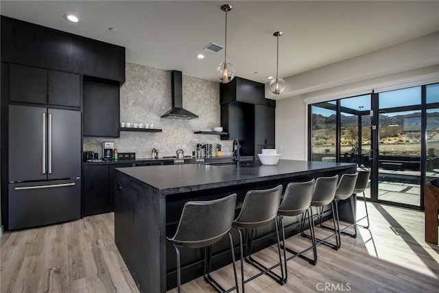 kitchen with wall chimney exhaust hood, a breakfast bar, paneled built in fridge, an island with sink, and pendant lighting