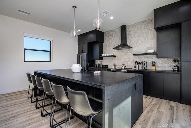 kitchen featuring tasteful backsplash, a kitchen island with sink, hanging light fixtures, wall chimney exhaust hood, and a breakfast bar