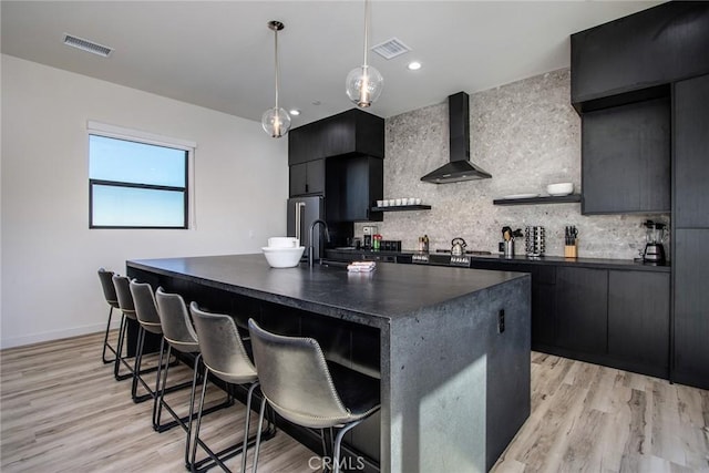 kitchen featuring decorative backsplash, decorative light fixtures, a kitchen island with sink, and wall chimney exhaust hood