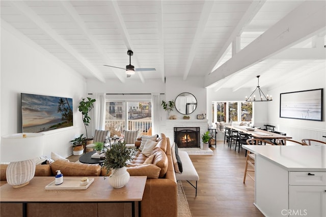 living room with ceiling fan with notable chandelier, light wood-type flooring, vaulted ceiling with beams, and a brick fireplace