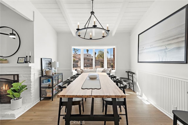 dining room featuring an inviting chandelier, beamed ceiling, light hardwood / wood-style floors, and a brick fireplace