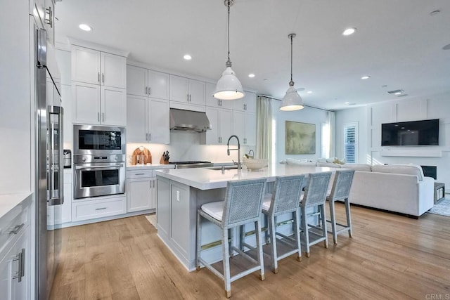 kitchen with sink, wall chimney range hood, decorative light fixtures, a center island with sink, and white cabinets