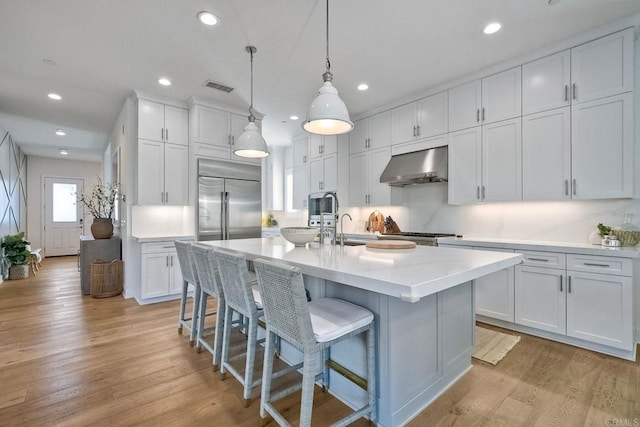 kitchen with pendant lighting, a kitchen island with sink, built in appliances, wall chimney exhaust hood, and white cabinetry