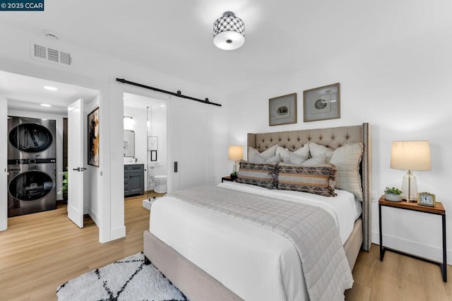 bedroom with a barn door, stacked washer / dryer, ensuite bath, and wood-type flooring