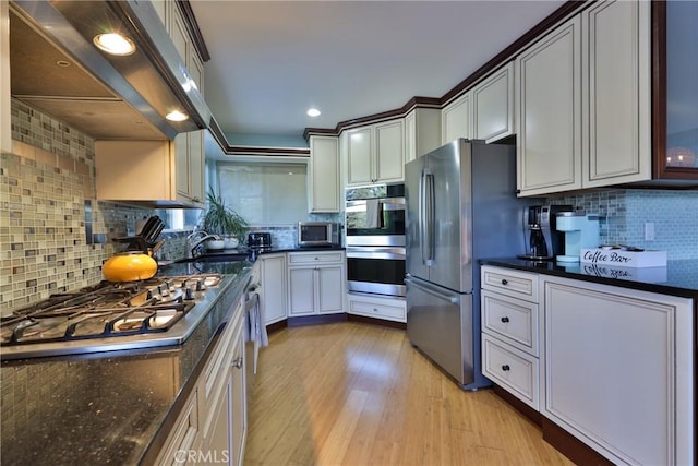 kitchen with appliances with stainless steel finishes, exhaust hood, dark stone counters, backsplash, and light wood-type flooring