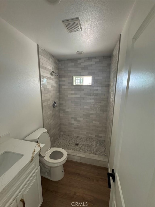 bathroom featuring toilet, wood-type flooring, tiled shower, vanity, and a textured ceiling