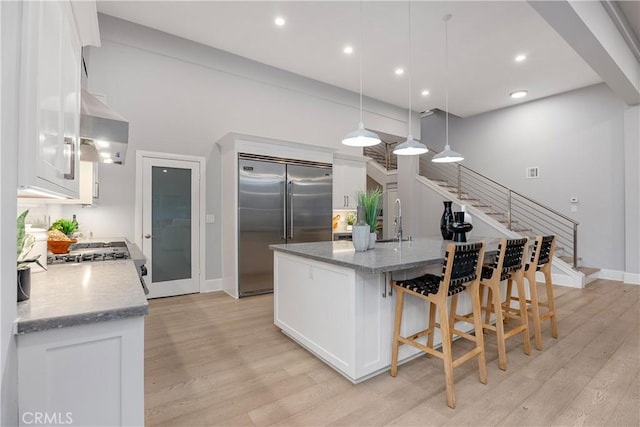 kitchen with stainless steel built in refrigerator, white cabinetry, light stone countertops, wall chimney range hood, and a center island with sink