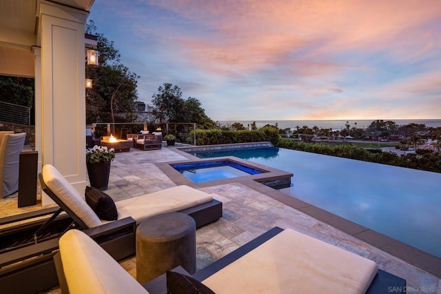 pool at dusk with an in ground hot tub, a water view, a patio, and a fire pit